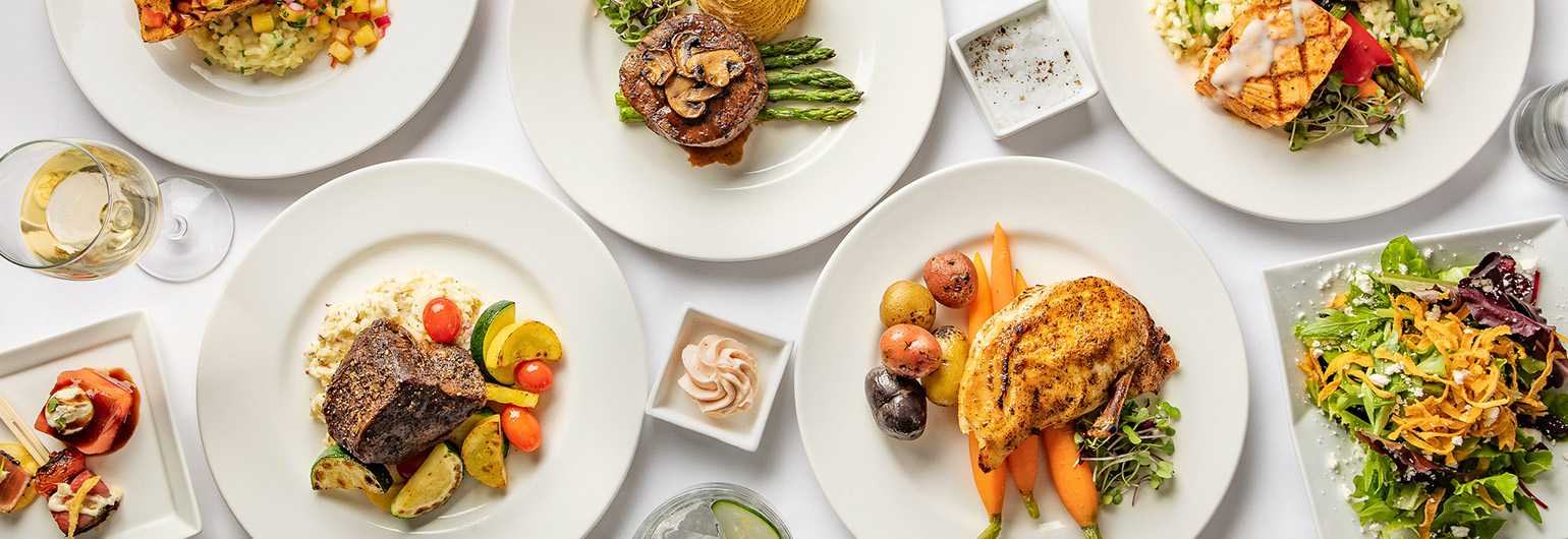 A variety of entrees displayed on a table with white plates including steak with asparagus, chicken and carrots, and more